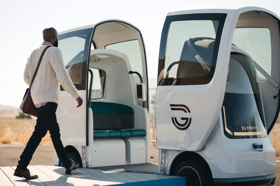 Man wearing sweater and messenger bag walking into white autonomous four-person Glydcar vehicle.